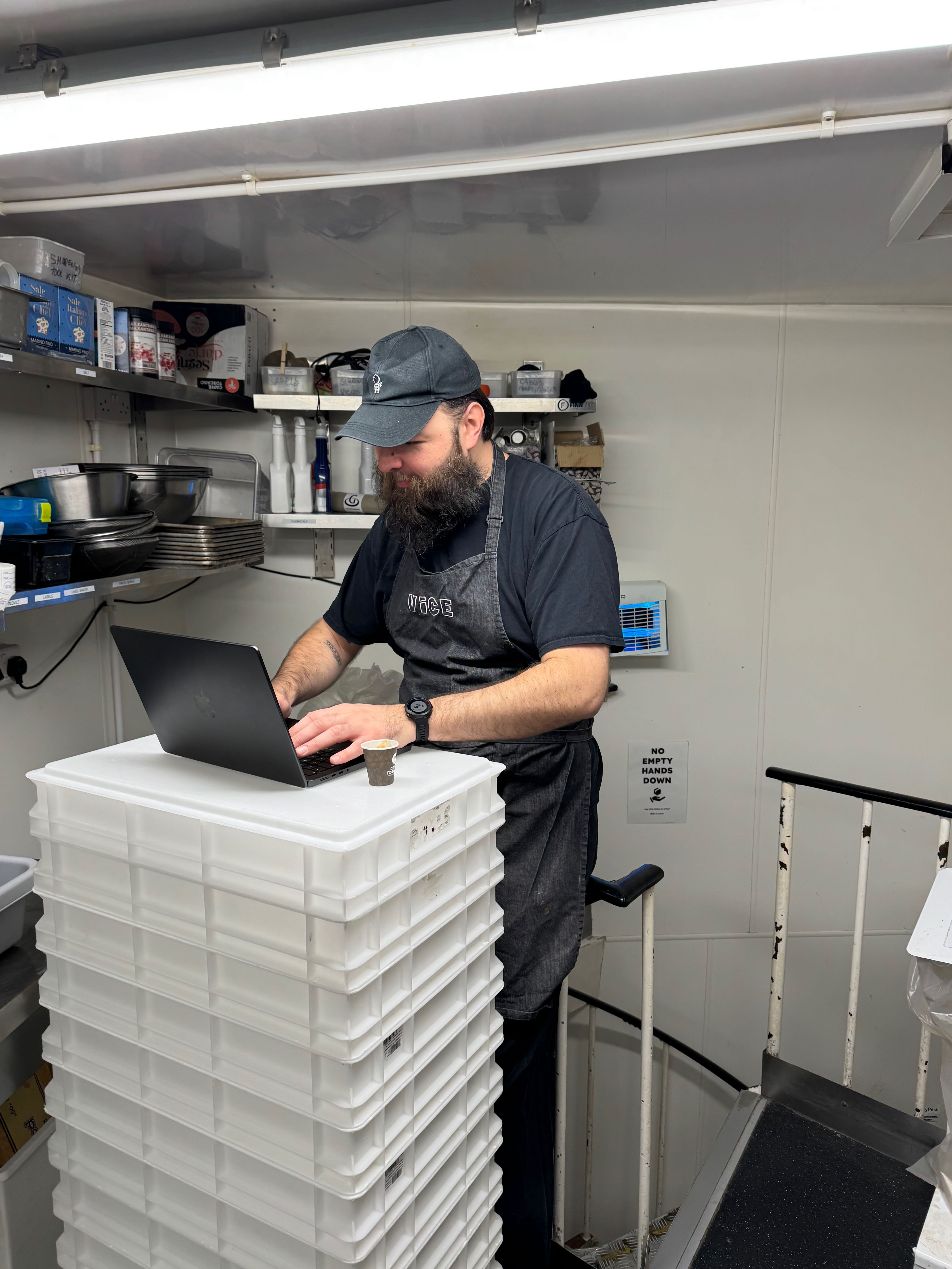 Allan in Vice Kitchen uniform working on a laptop in the kitchen prep area